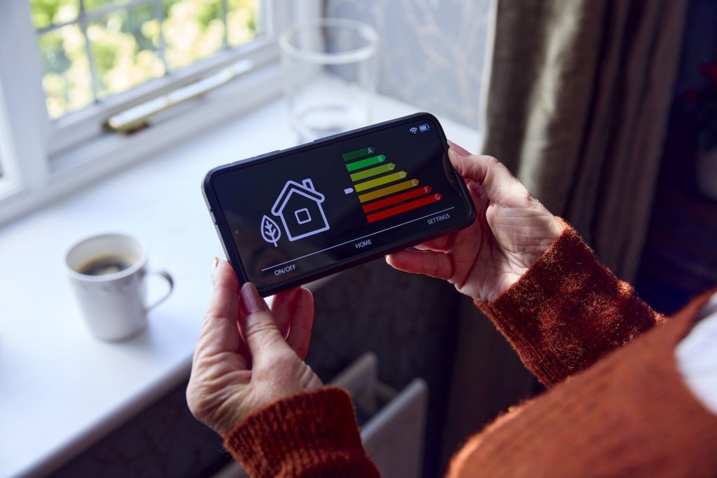 Senior Woman With Smart Meter Trying To Keep Warm By Radiator During Cost Of Living Energy Crisis